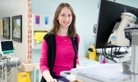 A nurse stands at a computer terminal and smiles at the camera