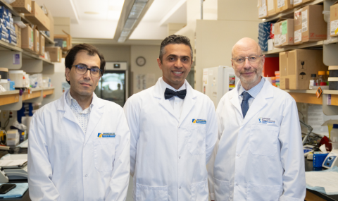 Three individuals wearing white lab coats stand side by side in a laboratory setting, facing the camera. The laboratory workspace includes shelves on both sides stocked with labeled boxes, containers, and scientific supplies, along with benchtop equipment and instruments. One individual at the centre is wearing a bow tie, while the others wear collared shirts. Visible logos on the lab coats indicate a research institute affiliation.