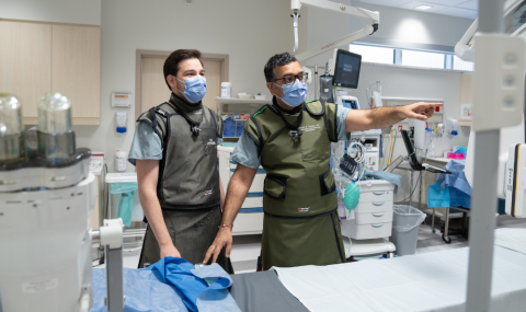 Two physicians stand in a clinical room dressed in protective equipment and point at a screen