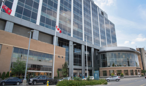 A large modern hospital building with a glass façade and multiple floors, reflecting clouds in its windows. In front of the building are several tall flagpoles displaying the Canadian flag and a red provincial flag. A curved entrance with glass walls sits to the right, with “B Entrance” signage visible. Cars are parked along the street in front, and small trees, shrubs, and a sidewalk line the foreground under a clear blue sky.