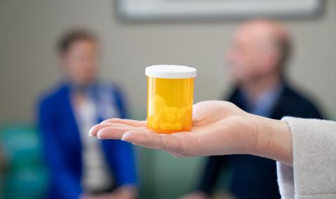 A hand holds a prescription bottle containing capsules, with two people blurred in the background during a research discussion.