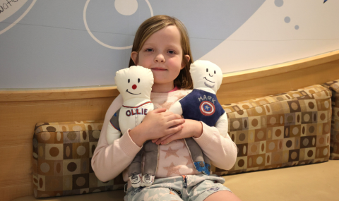 A young girl holds two stuffed dolls and smiles at the camera. 