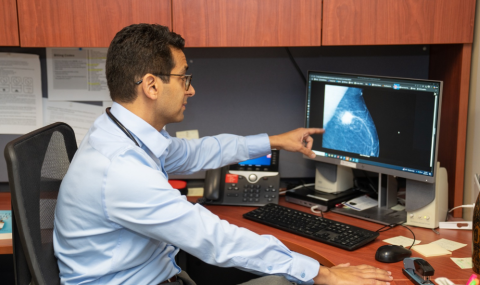 A physician sits at a desk, looking at his computer, which shows an image of breast cancer.