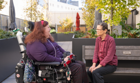 Martha in a motorized wheelchair wearing a dark purple shirt and dark purple plaid pants, sitting outside in the rooftop garden with Cathy, wearing a maroon shirt and black pants