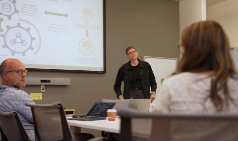 A researcher stands at the front of a meeting room presenting a slide about an AI-driven frailty score to colleagues seated around a table.