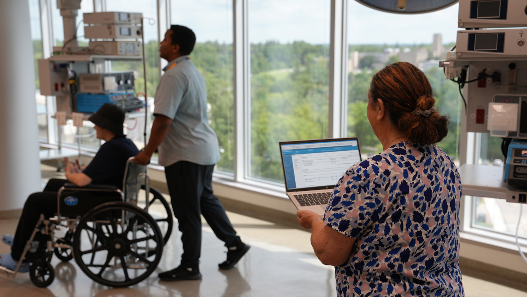 Nurse holding laptop while staff member pushes patient in a wheelchair 