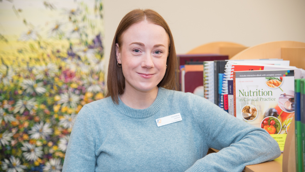 Registered Dietitian Christine Balsillie stands in the Health Sciences Library at Victoria Hospital. She's resting her left arm on a bookshelf holding a textbook called "Nutrition in Clinical Practice" and a colourful floral painting is behind her.