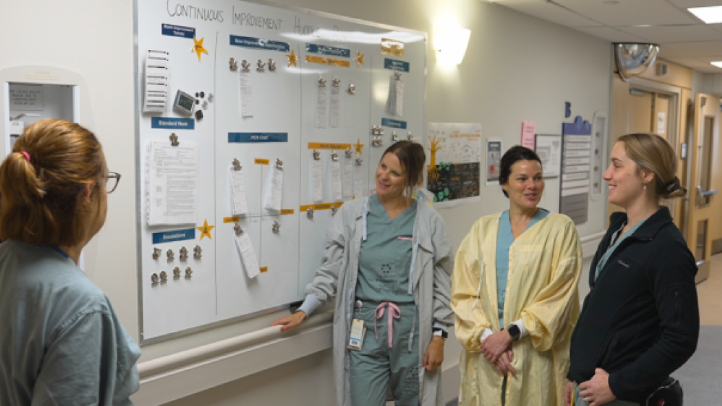 Four health-care professionals discuss information displayed on a whiteboard in a hospital corridor.