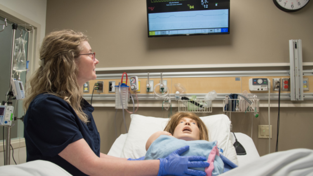 A healthcare professional in dark scrubs and gloves attends to a medical mannequin in a hospital bed. Medical equipment is mounted on the wall, and a monitor displays patient information.