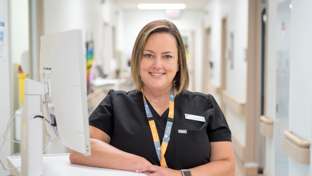 Jen Baker, wearing black scrubs, leaning up against counter with computer screen.