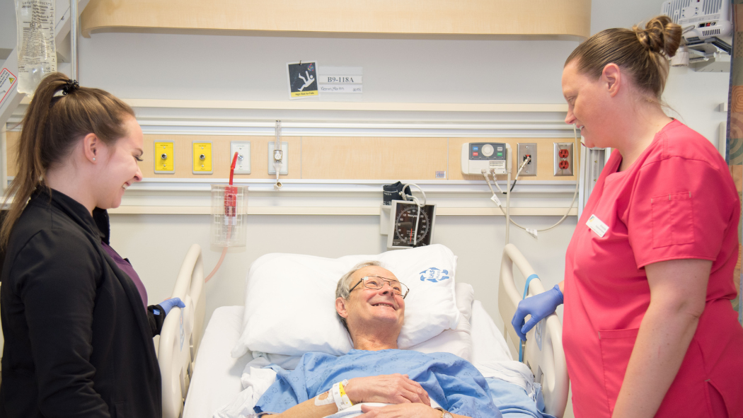 An elderly patient in a hospital bed smiles at two medical staff members, one in black and the other in red scrubs, who stand beside him.