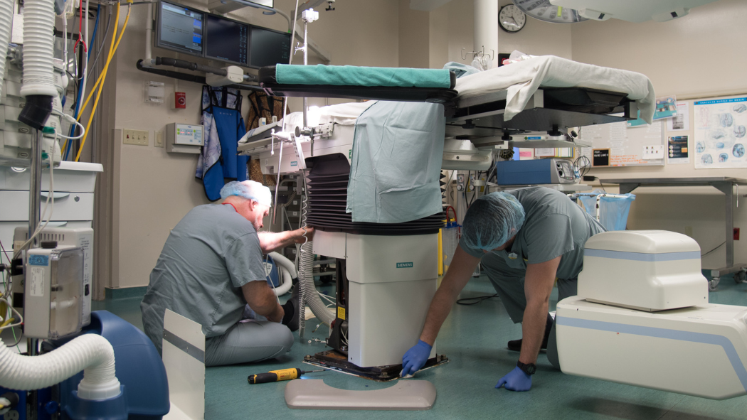 Two biomedical engineering technologists perform maintenance on a table in an operating room