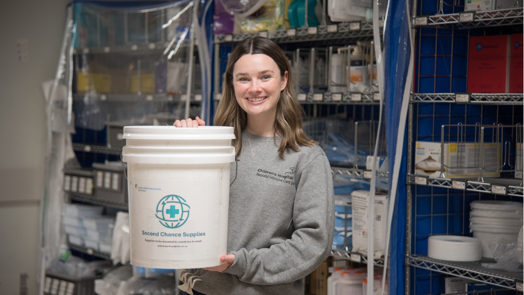 Person smiling and holding a white bucket labeled "Second Chance Supplies" in a storage room with shelves of medical supplies.