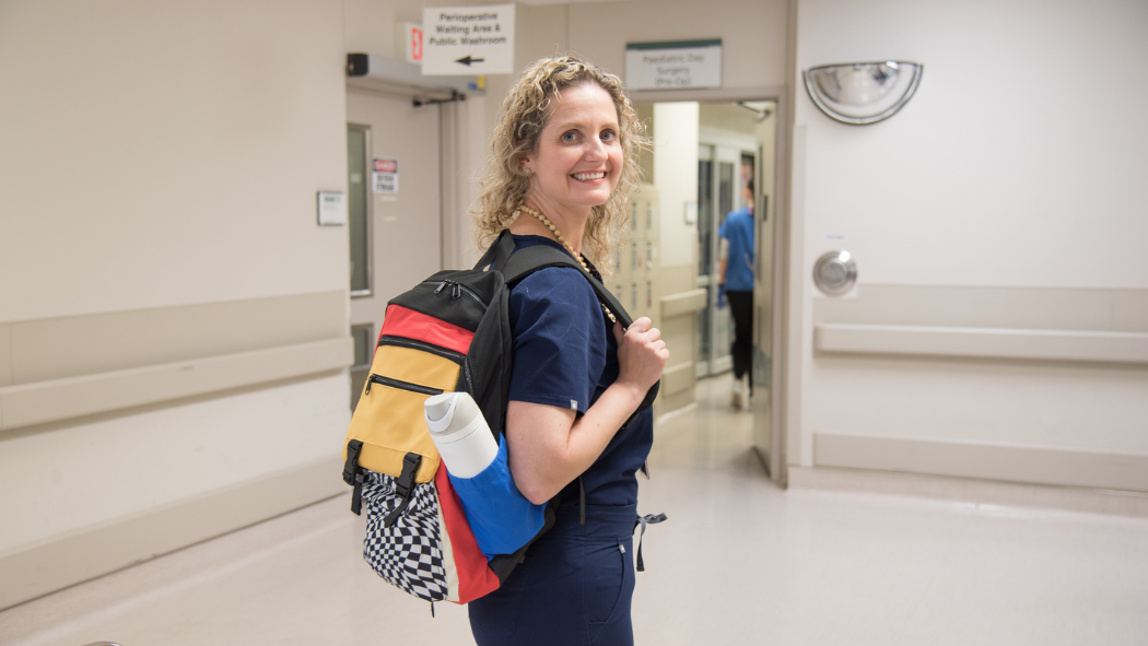 Smiling woman in scrubs stands in a hospital hallway with a colorful backpack over her shoulder, looking back at the camera.