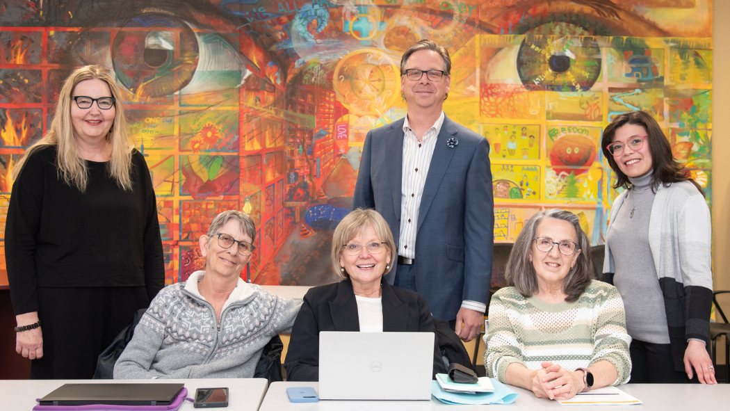 Seven adults pose for a group photo in a brightly lit room, seated at a table with laptops and tablets, in front of a colorful mural featuring large painted eyes.