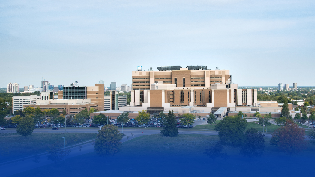 A large hospital complex with multiple buildings is shown, surrounded by trees and parking lots, with a cityscape visible in the background under a clear sky.