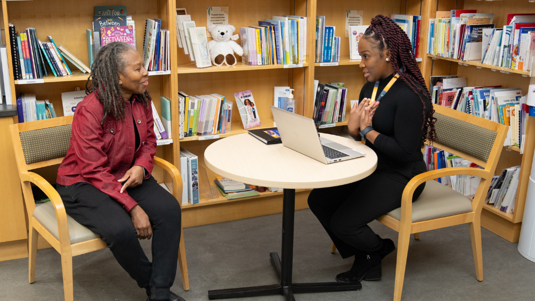 Two Black women talking while seated at a table in front of a bookshelf.