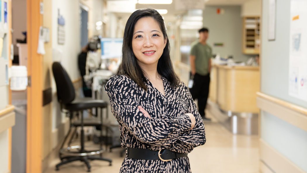 a woman stands with her arms crossed while smiling at the camera in a busy hospital hallways