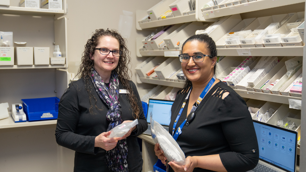 Female pharmacist holding pharmaceutical supplies smiles at camera standing next to female pharmacy resident who is also holding pharmaceutical supplies in a medication room