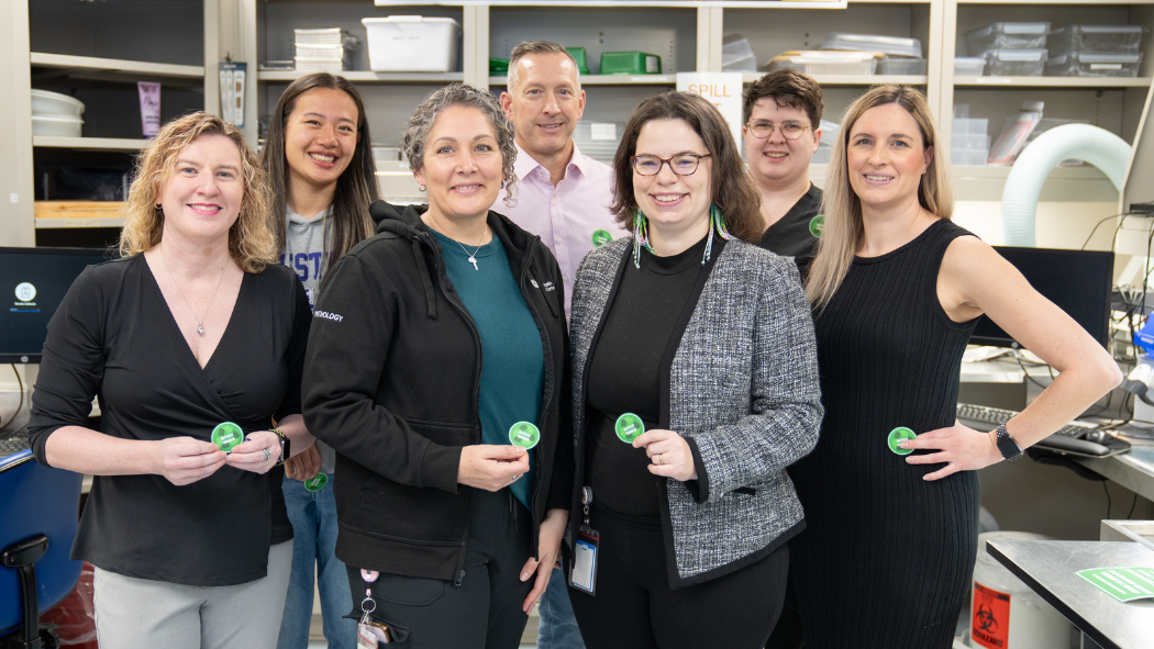 Seven people standing together in an office or lab setting, each holding a green circular badge and smiling at the camera.