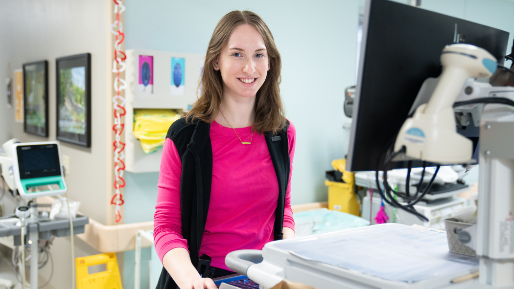 A nurse stands at a computer terminal and smiles at the camera