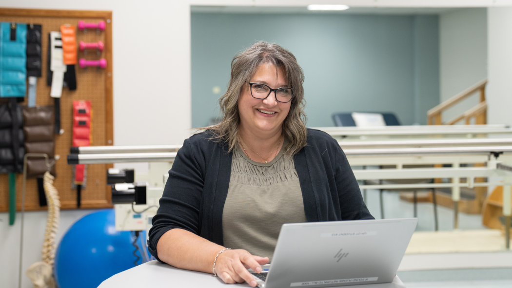 Patrice Green smiling sitting infront of laptop wearing an olive green shirt and black sweater
