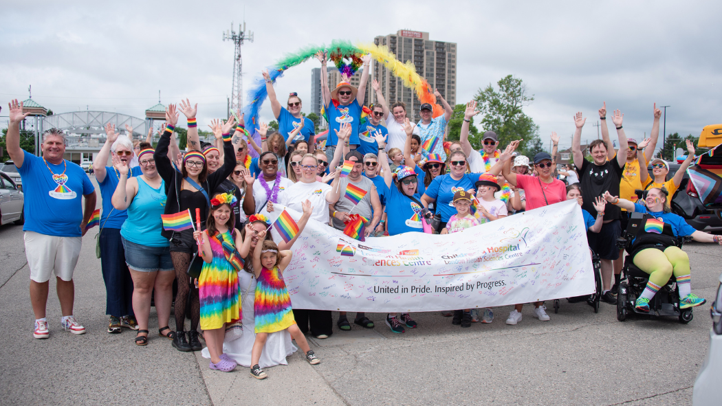 A large group of LHSC employees at the London Pride Parade in colourful clothing, standing behind a banner that reads "United in Pride. Inspired by Progress"