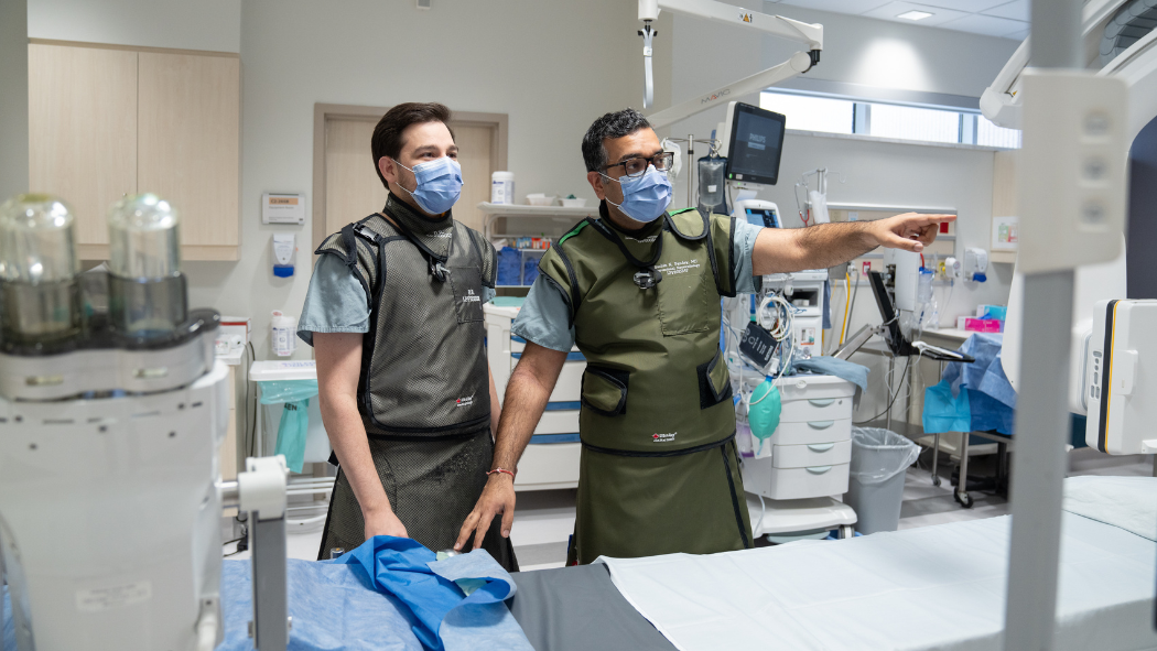 Two physicians stand in a clinical room dressed in protective equipment and point at a screen