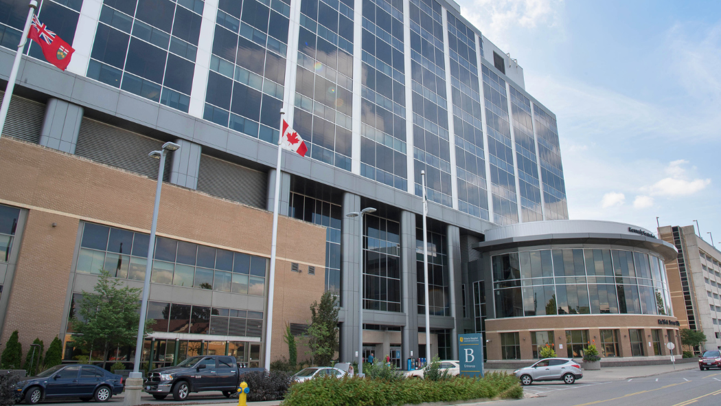 A large modern hospital building with a glass façade and multiple floors, reflecting clouds in its windows. In front of the building are several tall flagpoles displaying the Canadian flag and a red provincial flag. A curved entrance with glass walls sits to the right, with “B Entrance” signage visible. Cars are parked along the street in front, and small trees, shrubs, and a sidewalk line the foreground under a clear blue sky.