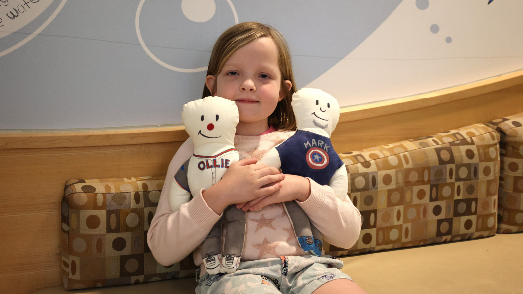 A young girl holds two stuffed dolls and smiles at the camera. 
