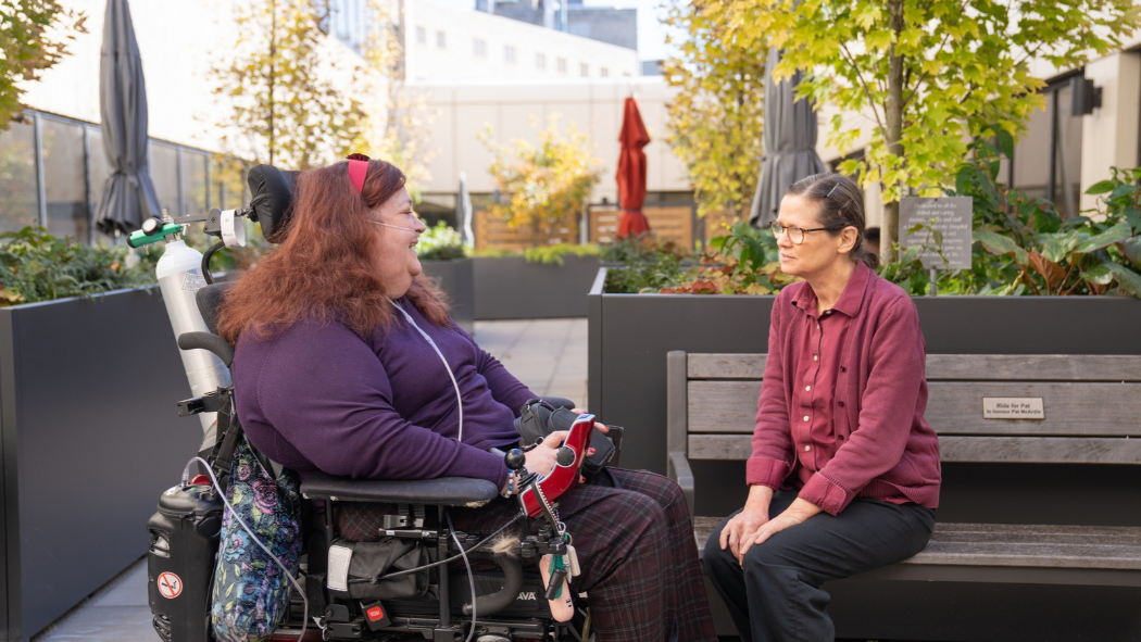Martha in a motorized wheelchair wearing a dark purple shirt and dark purple plaid pants, sitting outside in the rooftop garden with Cathy, wearing a maroon shirt and black pants